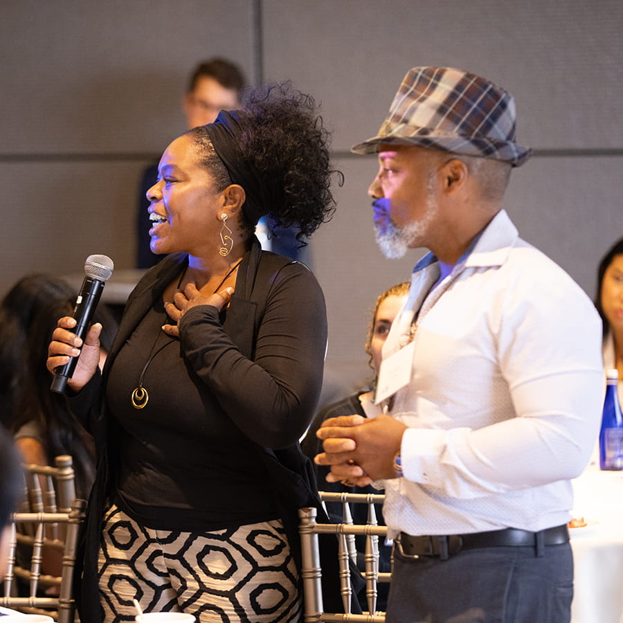 BEST Boston 2025 - woman standing in dress attire holding microphone with another attendee in another member in checkered porkpie hat and white casual business shirt (no tie)  looks on