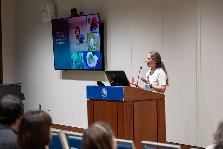 Juanita Urban-Rich stands at a podium in front of a crowd, giving her lecture. Behind her, a screen displays the words,